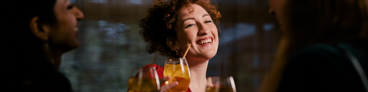 Een jonge vrouw kijkt lachend naar haar vrienden. Ze danst en heeft een cocktail in haar hand.